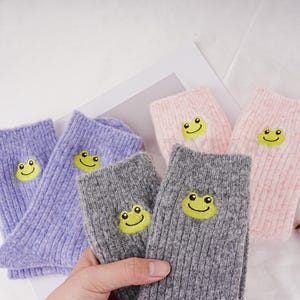 May include: Four pairs of cosy socks with a cute frog design. The socks are in purple, grey, and pink.