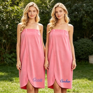 May include: Two pink spa wraps with a strapless design and button closure. The wraps have a wrap-around style with a split front. One wrap has the name "Scott" in blue script, and the other has "Amber". The background is a green, outdoor setting.