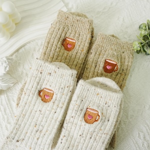 May include: Four pairs of cozy socks in cream and tan, each embroidered with a coffee cup design. The socks have a speckled texture and a ribbed knit pattern, perfect for a comfortable and warm feel.