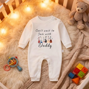 May include: White baby onesie with long sleeves and snap closures. The onesie features the text "Can't wait to Jam with Daddy" with colorful guitar graphics. A teddy bear and baby toys are in the background.