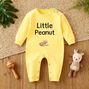 Puede incluir: Un body amarillo para bebé con mangas largas y cierres de botones a presión. La parte delantera presenta el texto "Little Peanut" sobre un gráfico de un cacahuete y dos mitades de cacahuete. Un sonajero de madera y un peluche están junto al body.