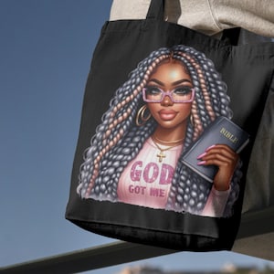 May include: Black tote bag featuring a colourful illustration of a woman with long braids wearing glasses and a pink shirt with the text "God Got Me". She is holding a book with the word "Bible" on the cover.