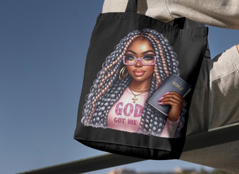 May include: Black tote bag featuring a colourful illustration of a woman with long braids wearing glasses and a pink shirt with the text "God Got Me". She is holding a book with the word "Bible" on the cover.