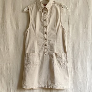 May include: A sleeveless collared dress in a light beige color. The dress has a button-up front, two patch pockets, and a fitted silhouette.