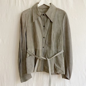 Könnte beinhalten: Eine graue Wildlederjacke mit Kragen, langen Ärmeln und einem Gürtel. Die Jacke hat einen Knopfverschluss und zwei Taschen.