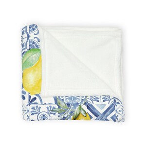 K&ouml;nnte beinhalten: Eine wei&szlig;e Fleece-Decke mit einem blauen und wei&szlig;en Fliesenmuster und gelben Zitronen. Die Decke ist in der H&auml;lfte gefaltet und das Fliesenmuster ist auf der Vorder- und R&uuml;ckseite sichtbar.