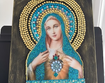 May include: A beaded artwork depicting the Virgin Mary with a heart on her chest. The heart is made of turquoise and gold beads. The artwork is framed in a dark brown frame and signed "Mayatt".