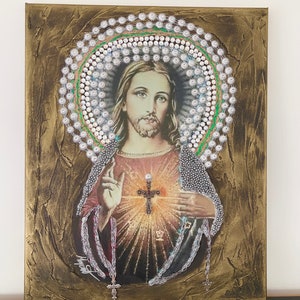 May include: A painting of Jesus Christ with a beaded halo and a cross on his chest. The painting is on a canvas with a gold background.