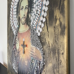 May include: A religious art piece featuring a portrait of Jesus Christ with a crown of thorns made of silver beads and a cross over his heart. The background is a textured gold canvas.
