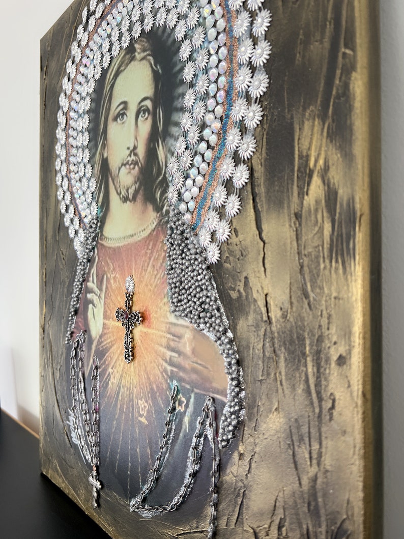 May include: A religious art piece featuring a portrait of Jesus Christ with a crown of thorns made of silver beads and a cross over his heart. The background is a textured gold canvas.