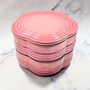 May include: A pink ceramic container, flower-shaped, with a lid and base. The edges are scalloped. The Le Creuset logo is on the lid. The container is stacked. It sits on a white marble surface. The container is suitable for kitchen use.