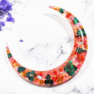 May include: A crescent moon-shaped resin tray with a glittery orange base and green and gold accents. The tray is decorated with embedded green and gold glitter and has a smooth, glossy finish.