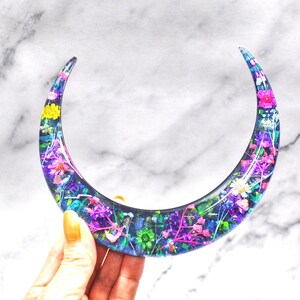 May include: A crescent moon-shaped resin ornament with embedded pressed flowers in shades of pink, purple, blue, and yellow.