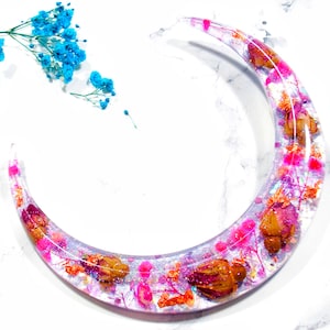 May include: A crescent moon-shaped resin tray with pink, orange, and white dried flowers and glitter embedded inside.