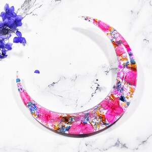 May include: A crescent moon shaped resin ornament with pink and gold glitter embedded in the resin. The resin is filled with dried pink flowers and gold leaf.