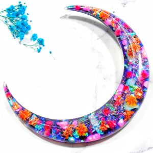 May include: A crescent moon-shaped resin ornament with a dark blue background and embedded colourful glitter, small flowers, and iridescent flakes.