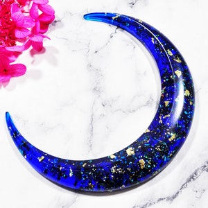 May include: A crescent-shaped hair accessory in a deep blue hue, embedded with gold flakes. The curved barrette is set against a white marble surface, with bright pink flowers in the background. A unique hair clip.
