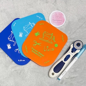 Könnte beinhalten: Drei orangefarbene, blaue und hellblaue Plastik-Eckenmacher-Schablonen mit Maßen in Zentimetern. Die Schablonen zeigen eine Nähmaschine, Garn, eine Schere, Stecknadeln und Knöpfe. Ein blauer Rollschneider und ein weißer Stift sind ebenfalls enthalten.