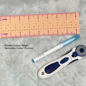 Op de afbeelding: Een beige liniaal met fuchsia markeringen en perforaties, een witte Cricut pen met een blauwe punt en een wit en blauw roterend mes. De liniaal is gemarkeerd in inches.