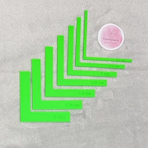 Può includere: Un set di nove righelli d'angolo in plastica verde di diverse dimensioni, che vanno da 0,5 cm a 2 cm. I righelli sono disposti a forma di piramide, con il righello più grande in basso e il più piccolo in alto.