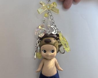 Puede incluir: Un llavero con una pequeña muñeca que lleva pantalones cortos azules y un sombrero marrón con orejas de oso. El llavero tiene un cierre en forma de estrella y está adornado con lazos, cuentas y abalorios amarillos.