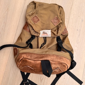 May include: A tan Kelty backpack with brown leather accents. The backpack has black straps and buckles, with a small white tag that reads "KELTY". The bag has a vintage, outdoorsy aesthetic.