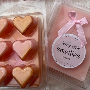 May include: A set of six heart-shaped wax melts in a clear plastic mold. The melts are a light pink color with a gold shimmer. The wax melts are packaged in a clear plastic box with a pink ribbon and a label that says "lovely little smellies wax co."