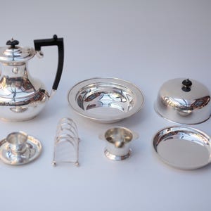 Puede incluir: Conjunto de piezas de servicio de plata antiguas, que incluyen una jarra con asa negra, cuencos, un plato cubierto, una taza y platillo pequeños, un tostador y un cuenco pequeño. Sobre una superficie blanca.