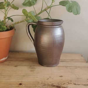 May include: A brown ceramic pitcher with a white handle. The pitcher is sitting on a wooden surface. A green plant is in the background.