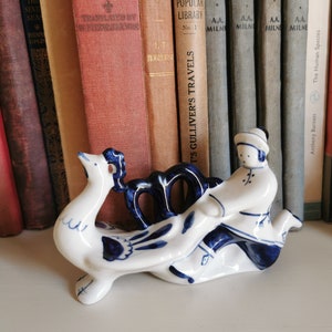 May include: A blue and white ceramic figurine of a person riding a rooster. The figurine is hand-painted with a traditional folk art style.