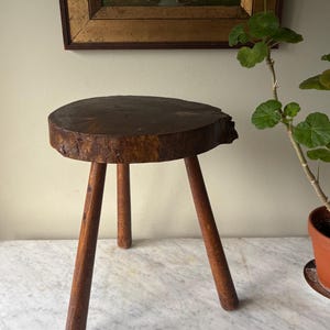 May include: A dark brown, three-legged wooden stool with a circular seat. The legs are angled and taper towards the floor. The stool is made of natural wood, showcasing the grain and texture. The stool is a decorative piece.