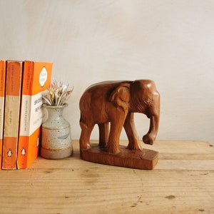 May include: A wooden elephant figurine with a natural brown finish. The elephant is standing with its trunk curled up. The figurine is on a wooden surface with a few books and a small vase with dried flowers.