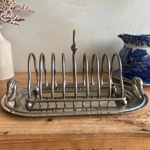 May include: Silver-toned metal toast rack with a swan design on each end. The rack has a handle and slots for toast. The oval base and vertical bars create a classic, vintage aesthetic. A decorative piece for the kitchen or dining room.