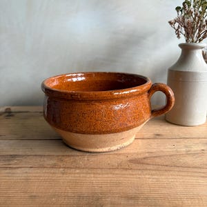 May include: A glazed ceramic bowl with a handle, featuring a rich brown upper section and a lighter, unglazed base. The bowl is displayed on a wooden surface. A small white vase with dried flowers is in the background.