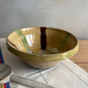 May include: A rustic, hand-painted ceramic bowl with a yellow interior and green and brown stripes. The bowl sits on a white and green striped cloth, with a wooden spoon in the foreground. The bowl has a diameter of approximately 20 cm.