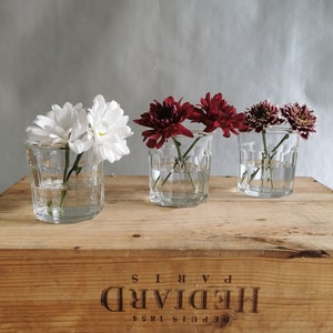 May include: Three glass bud vases with white and red flowers. The vases are clear glass with a textured design. The vases are sitting on a wooden crate with the text "Depuis 1854 HEDDIARD PARIS" printed on the front.