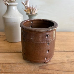 May include: A brown ceramic pot with a cylindrical shape and small feet. The pot has several small holes around the sides and a slightly flared rim. Dried flowers are in the background in a white and green vase.