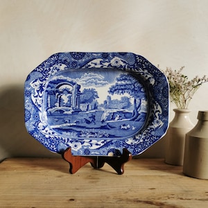May include: A blue and white ceramic serving platter with a floral design and a scene of a countryside landscape. The platter is oval shaped and has a raised rim.