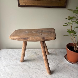 May include: A vintage wooden stool with a rectangular seat and three legs. The stool is made from light-coloured wood, showing the natural grain. The legs are angled outwards. The stool has a rustic, handcrafted appearance, suggesting it is an antique or vintage item.