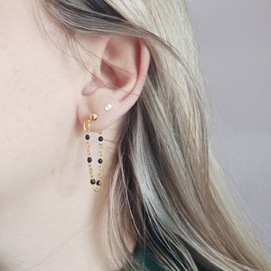 Könnte beinhalten: Goldfarbene Kettenohrringe mit schwarzen Perlen. Die Ohrringe sind an einem kleinen goldenen Kugelstecker befestigt. Ein kleiner, silberner, blütenförmiger Ohrstecker ist ebenfalls sichtbar. Die Person trägt einen dunkelgrünen Rollkragenpullover.