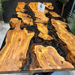 Könnte beinhalten: Ein großer Esstisch mit einer natürlichen Holzkante und einer schwarzen und orangefarbenen Epoxidharzeinlage. Der Tisch hat eine natürliche Holzoptik und ein schwarzes, X-förmiges Metallgestell. Der Tisch ist mit "Olive Wood" beschriftet.