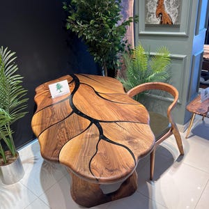 May include: A unique wooden table with a leaf-like design, featuring dark epoxy resin accents. The table is paired with a wooden chair with a green cushion. The table has the text "BRAVEWOOD" on it.