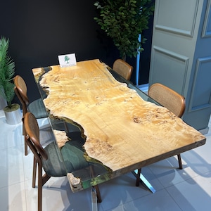 May include: A large wooden dining table with a live edge top and a clear epoxy resin fill. The table has a natural wood finish and is supported by a metal base. The table is surrounded by four chairs with woven seats.