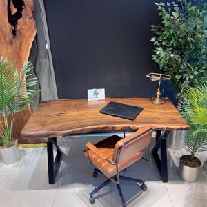 Puede incluir: Un escritorio de madera con borde vivo y patas de metal negro, una silla de oficina de cuero marrón y una lámpara de escritorio de estilo vintage. Una computadora portátil está sobre el escritorio. El escritorio está rodeado de plantas en macetas y una pared azul oscuro.