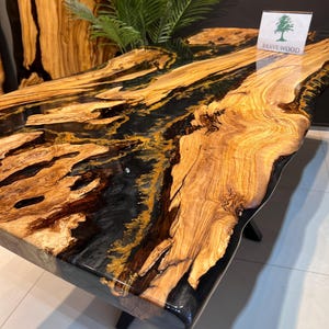 Könnte beinhalten: Eine große, naturbelassene Holztischplatte mit schwarzem und goldenem Epoxidharz. Die Tischplatte hat ein natürliches, rustikales Aussehen mit einer glatten, polierten Oberfläche. Das Epoxidharz erzeugt ein einzigartiges und auffälliges Design. Die Tischplatte wird von Brave Wood hergestellt.