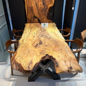May include: A large, natural wood dining table with a live edge top and a black metal X-shaped base. The table is surrounded by four chairs. The table top has a unique grain pattern and is made of a light wood with dark brown streaks. The table is labeled "Brave Wood".