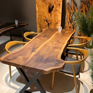 Puede incluir: Una mesa de comedor de madera grande con una tapa de borde vivo y una base en forma de X de metal negro. La mesa está rodeada por cuatro sillas con marcos de madera clara y asientos de cuero negro.