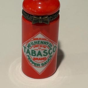 May include: A small, red Tabasco sauce bottle with a green neck and red cap. The label displays "TABASCO" and "McIlhenny Co. Avery Island, LA." A decorative bow is around the middle of the bottle. The bottle is a miniature size.