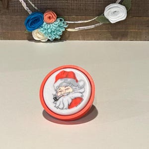 Puede incluir: Un botón redondo con borde rojo que presenta una ilustración de Papá Noel sonriendo. El botón está frente a un fondo de madera con una corona floral y la palabra "hello" en escritura blanca.