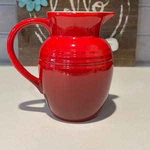 May include: A red ceramic pitcher with a handle. The pitcher has a smooth, glossy finish and a rounded shape.